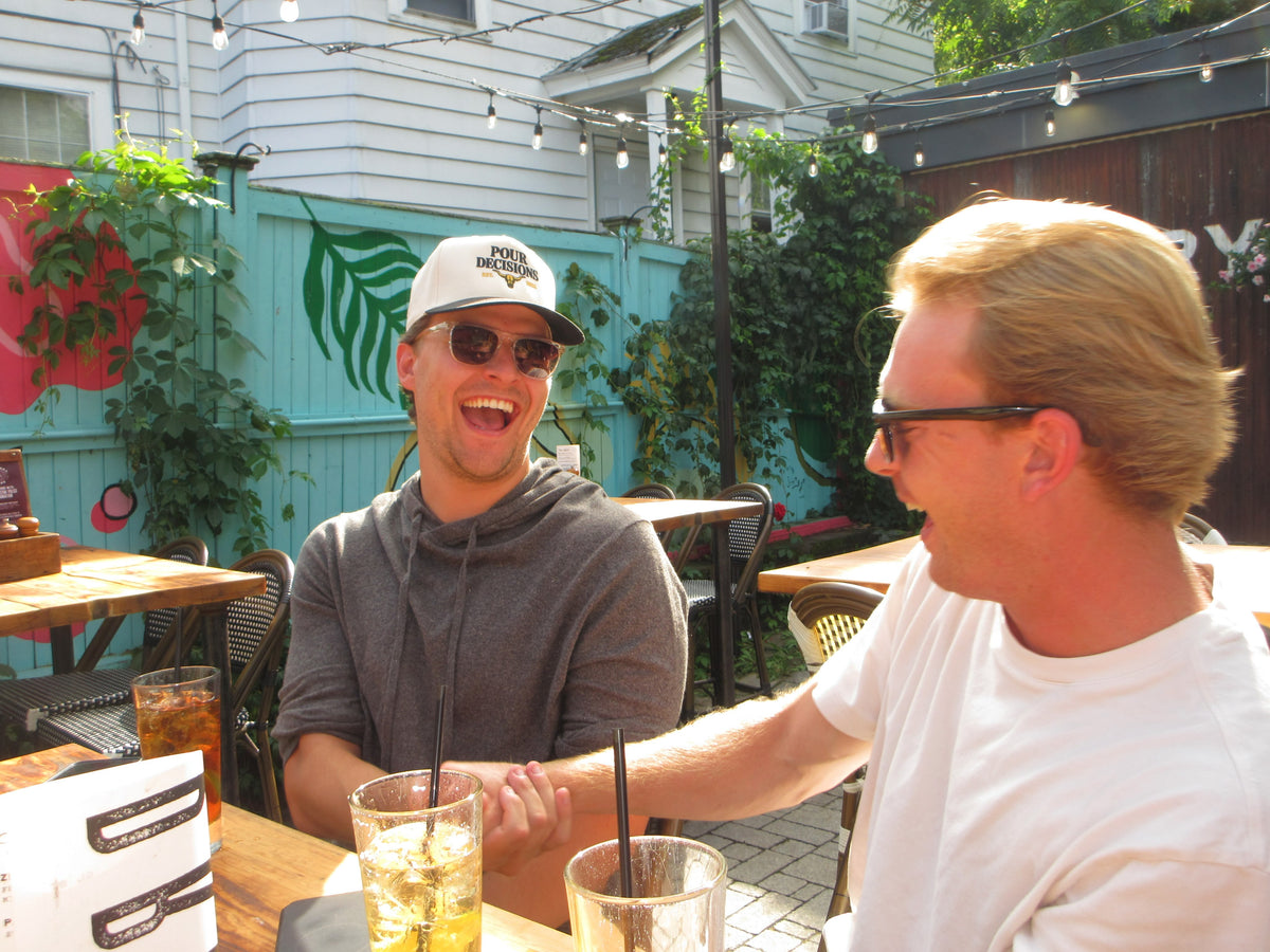 Two friends laughing on a bar patio, one in the Pour Decisions SnapBack